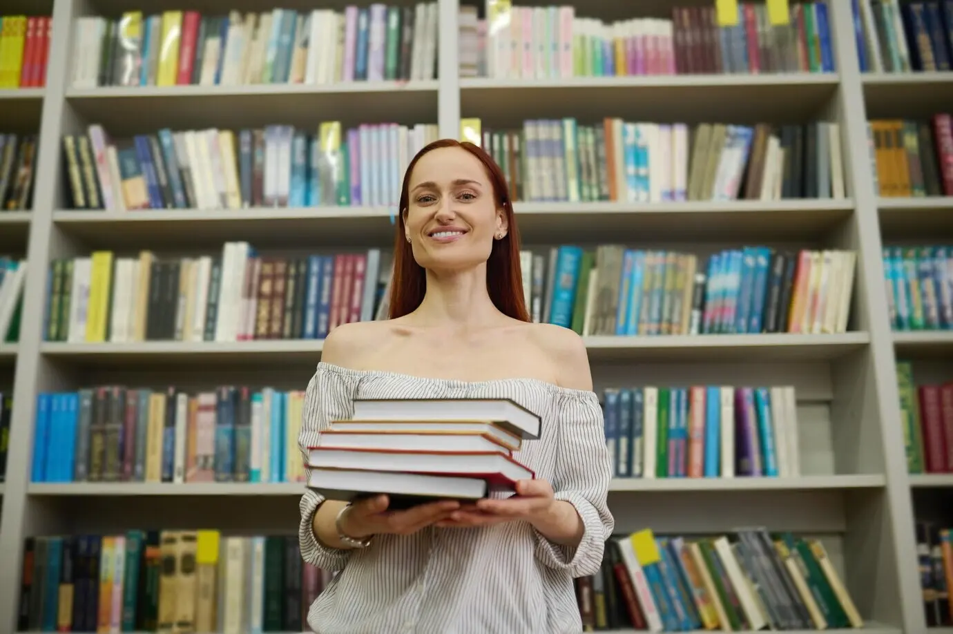 A woman holds a stack of books next to bookshelves.